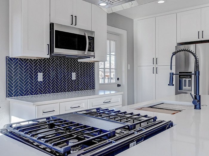 Modern white-cabinet kitchen with a stainless hood, gas cooktop on a white quartz island, and a deep-blue herringbone tile backsplash